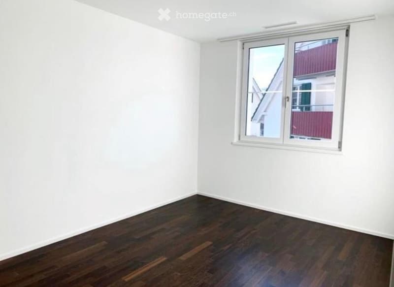 Empty room with white walls and dark wooden flooring, featuring a large window with white frame and blinds.