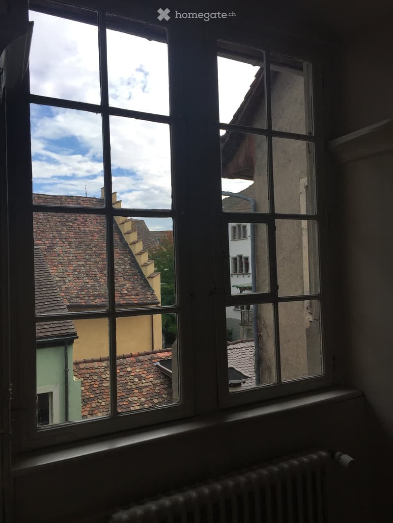 a window that looks out at a street scene, a nearby building, and a balcony with green railing