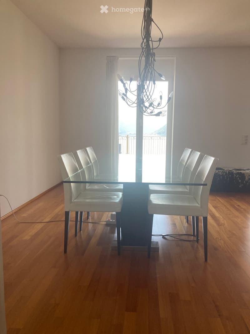 Dining room with a glass table and four white chairs, wooden flooring, and a chandelier hanging from the ceiling.