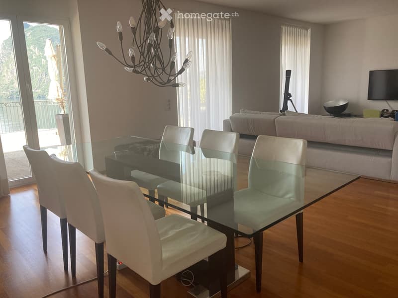 Modern living and dining area with a glass dining table, white chairs, a grey sofa, a chandelier, and a television. Large windows offer a scenic view.