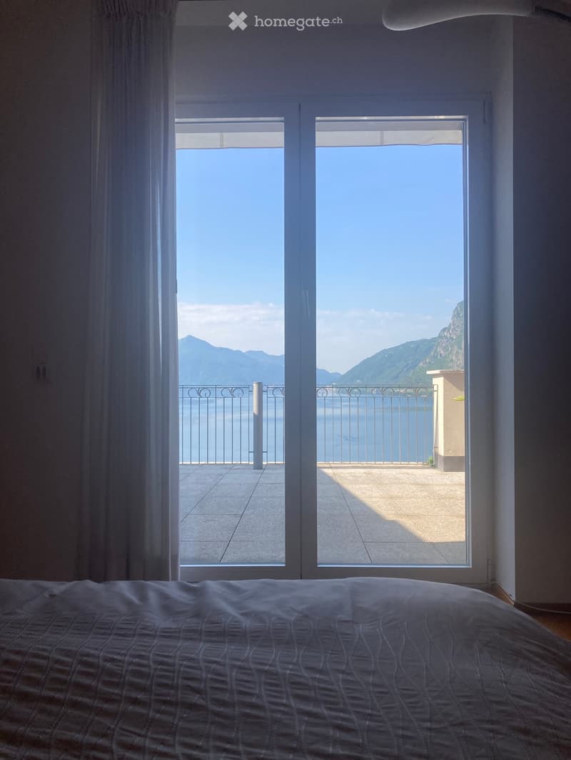 Bright bedroom with large windows opening to a balcony overlooking a scenic lake and mountains.
