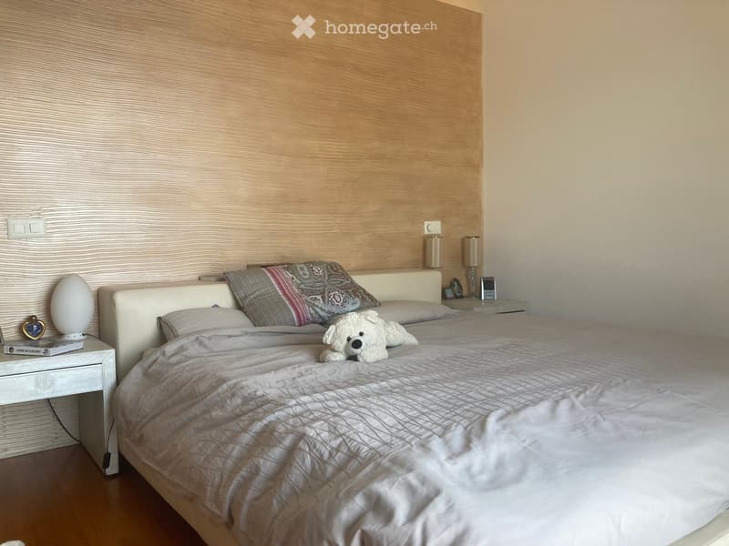Modern bedroom with a large bed, decorative pillows, a teddy bear, bedside tables with lamps, and a decorative egg-shaped lamp on a nightstand.
