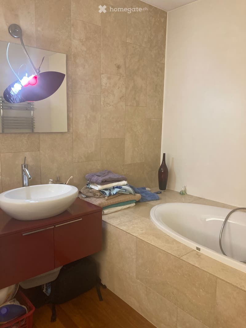 Modern bathroom with a white sink, bathtub, mirror, and neatly stacked towels on a shelf. A decorative vase is placed on the counter.