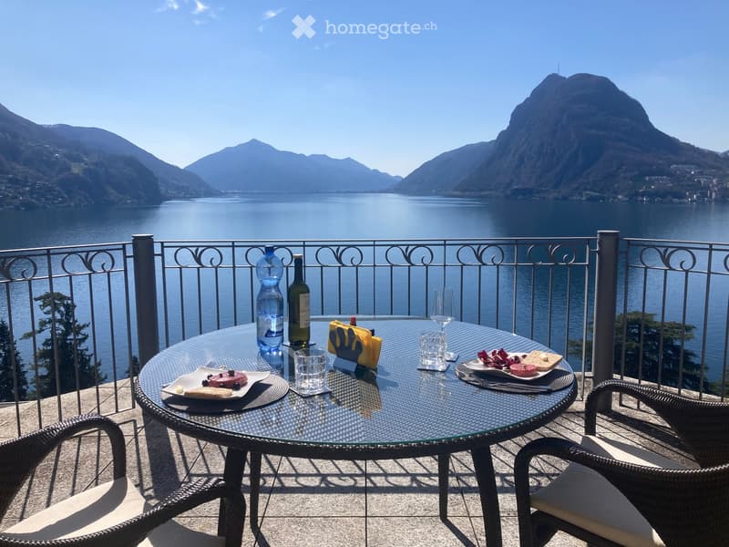 Balcony with a round glass table, two wicker chairs, and a scenic lake and mountain view.