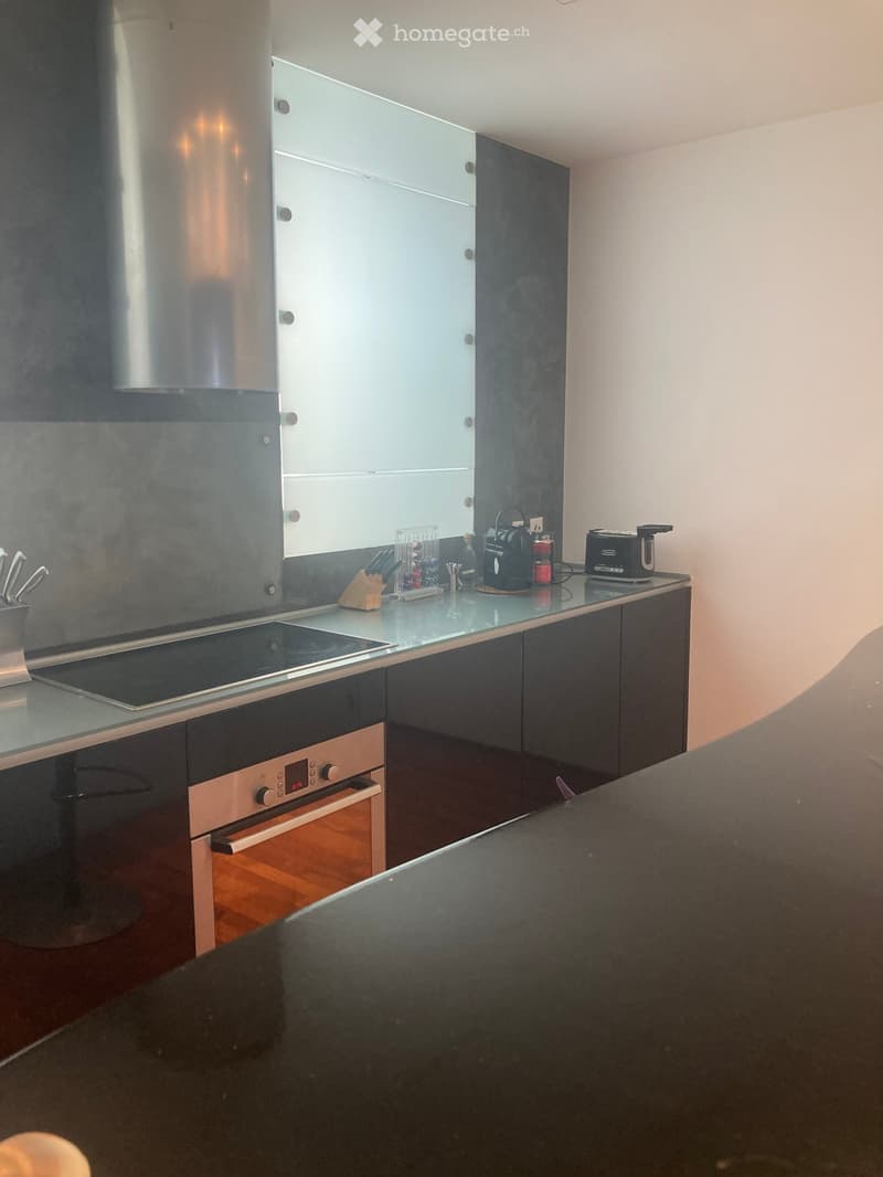 Modern kitchen with dark grey cabinets, a central island, and stainless steel appliances including an induction stove and oven. The countertop features a coffee maker, toaster, and knife block.