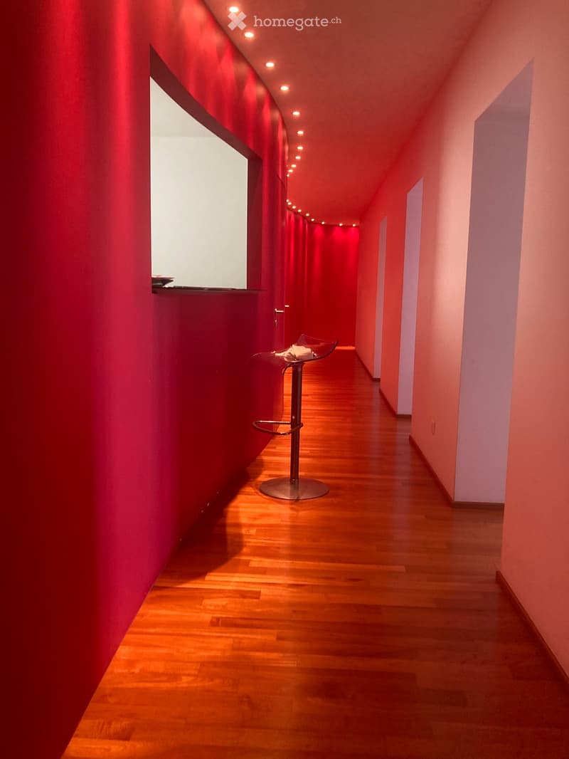 A narrow hallway with wooden flooring, red accent wall, and a single modern stool in the center.
