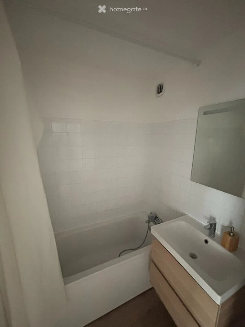 Bathroom with a bathtub, sink, mirror, and wooden cabinet. The walls are tiled, and there is a small window providing natural light.