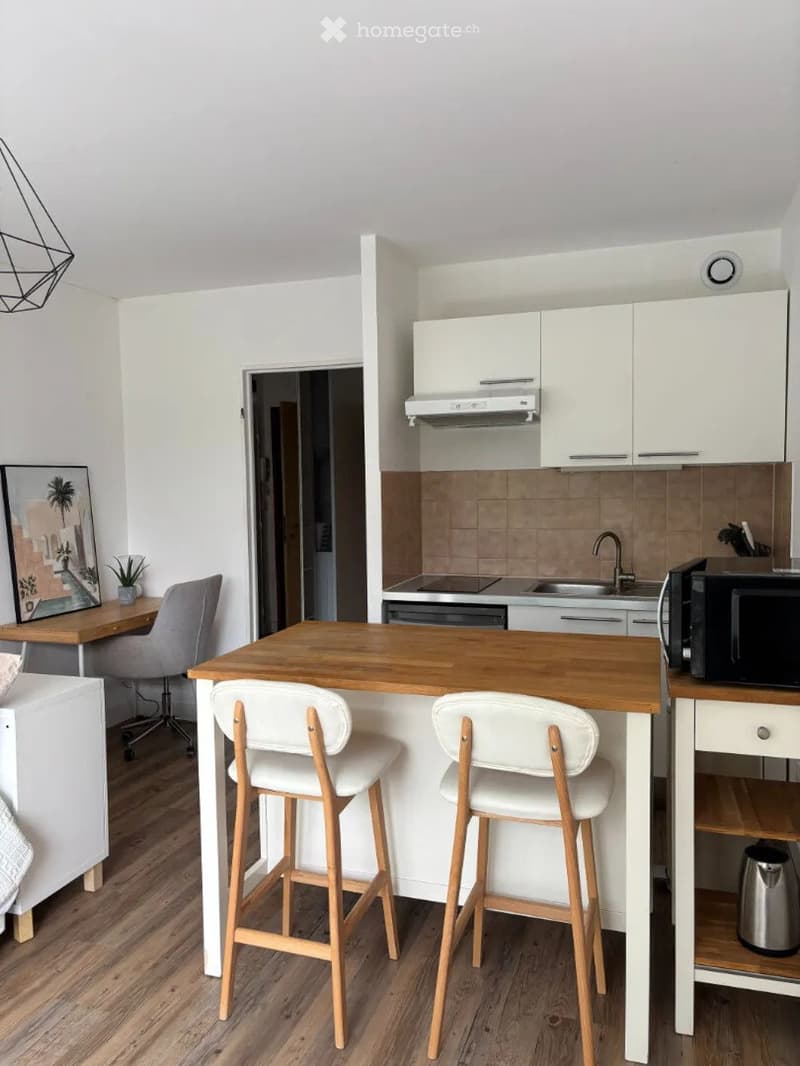 Modern kitchen with white cabinets, wooden island, bar stools, microwave, and a small desk with a chair and plant in the background.