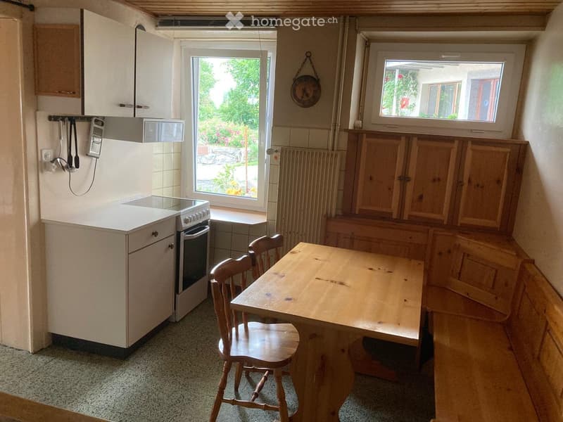 A small kitchen with white cabinets, a refrigerator, an oven, a wooden dining table, and chairs. There are windows with a view of greenery outside.
