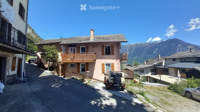 A two-story residential building with a wooden balcony, situated in a picturesque mountain village with clear blue skies and surrounding mountains.