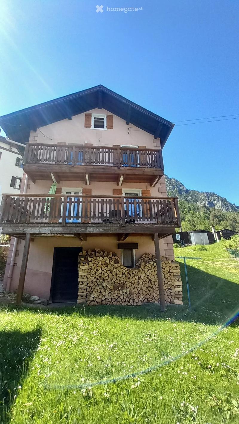 Two-story house with wooden balconies, light pink exterior, and a stack of firewood near the entrance. Surrounded by green grass and a mountain view in the background.