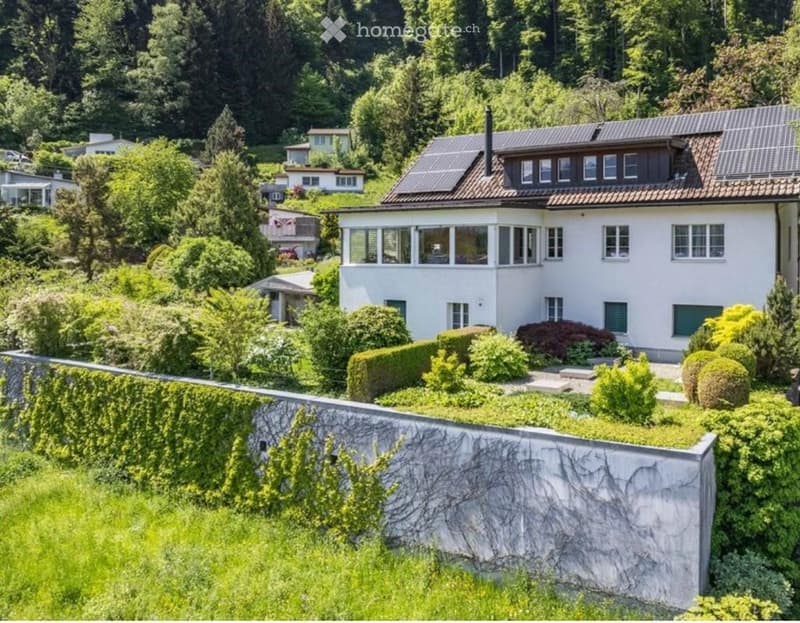 A two-story white house with a red-tiled roof, surrounded by lush greenery and trees. The house features multiple windows and a balcony area on the upper floor.