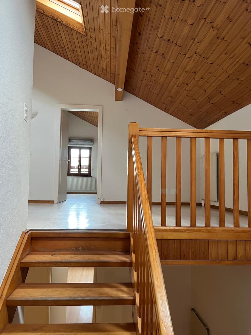 Wooden staircase with a wooden railing and a skylight above, leading to a bright, open space with white walls and a large window at the end.