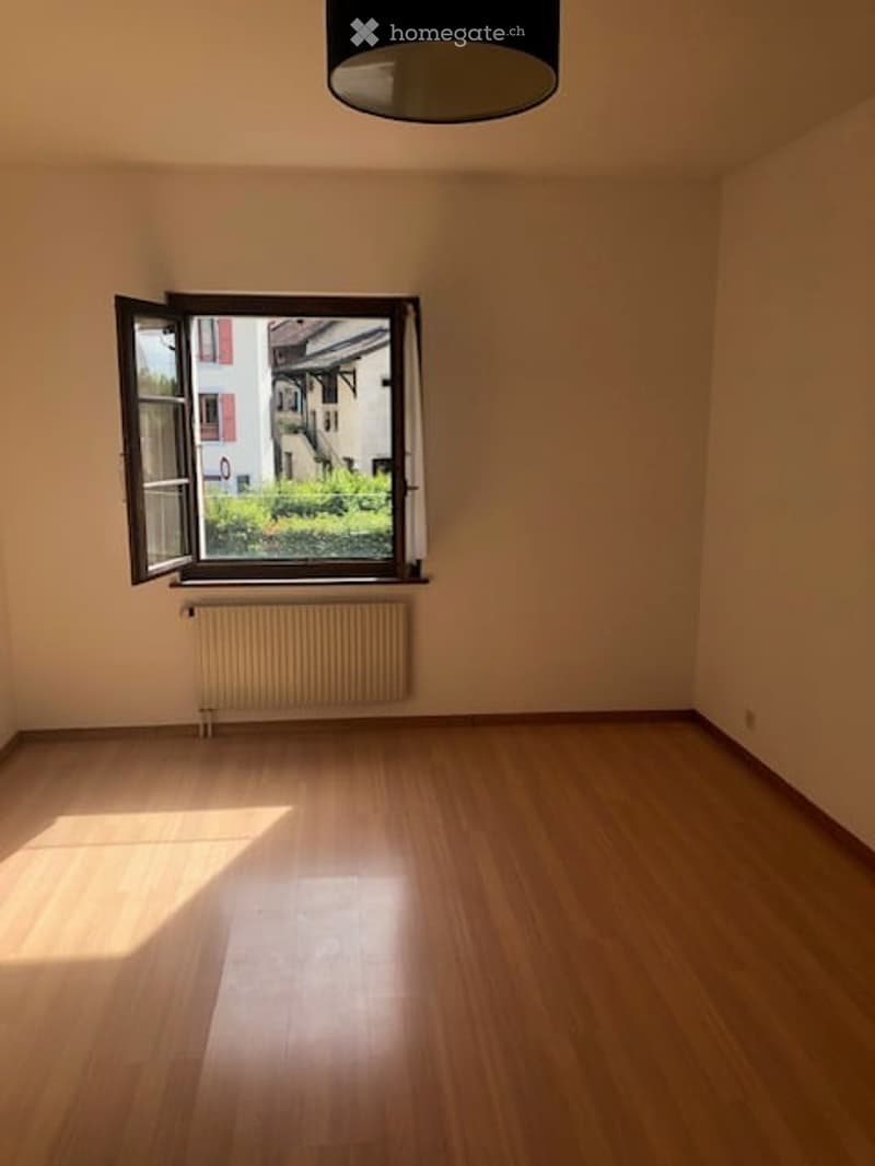 Empty room with wooden flooring, white walls, a radiator, and a large window with a view of a garden and neighboring building.
