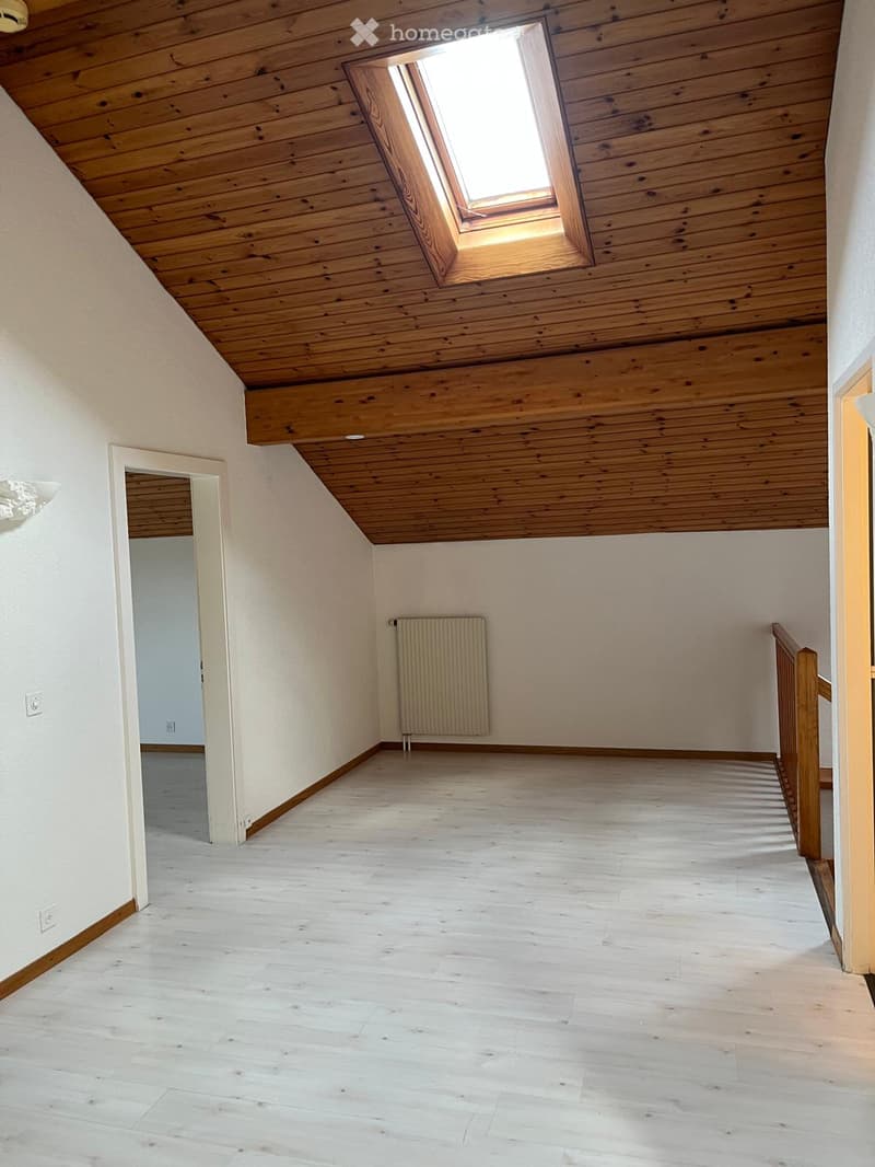 Empty room with wooden flooring, white walls, and a skylight window in the ceiling. A radiator is visible on one wall, and a doorway leads to another room.