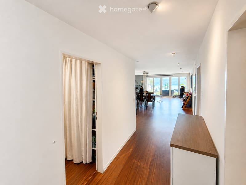 A bright, modern hallway with wooden flooring, white walls, and recessed lighting. A small wooden console table is positioned against the right wall, and a closet with beige curtains is visible on the left. The hallway leads to a dining area with chairs a