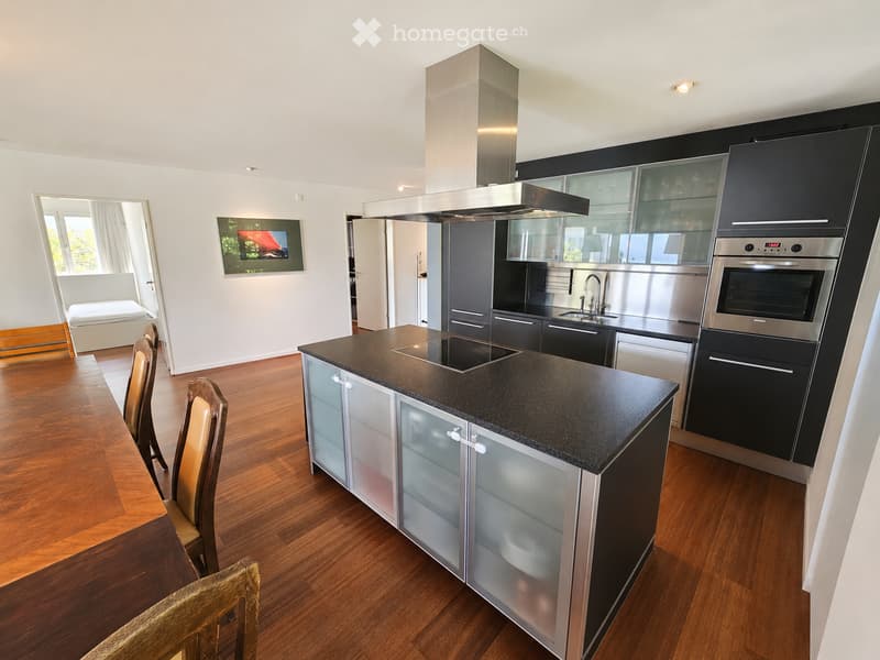 Modern kitchen with dark countertops, stainless steel appliances, and wooden flooring. Features a central island with a built-in stove, a sink with a modern faucet, and a range hood. There are four wooden chairs around a small table, and a picture frame i
