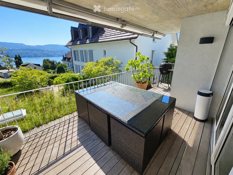 Balcony with wooden decking, glass-topped table, wicker chairs, barbecue, potted plants, and scenic mountain view.