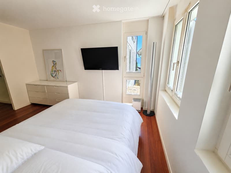 Modern bedroom with a large bed, white bedding, wooden flooring, a wall-mounted TV, a dresser, and a floor lamp near the window.