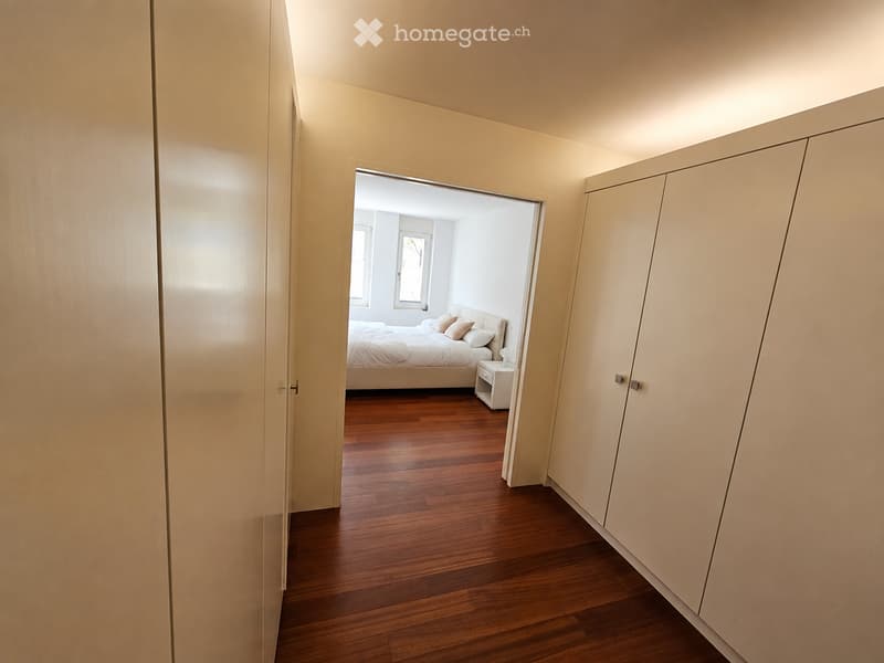 A hallway leads to a furnished bedroom with a wooden floor, a bed with pillows, and a nightstand.