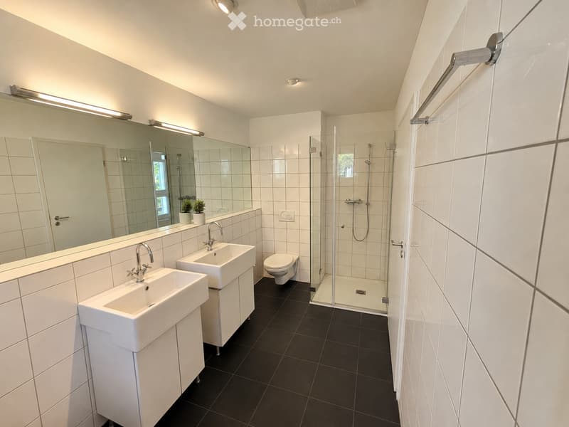 Modern bathroom with white tiles, two sinks, a toilet, a glass-enclosed shower, and a large mirror with integrated lighting.