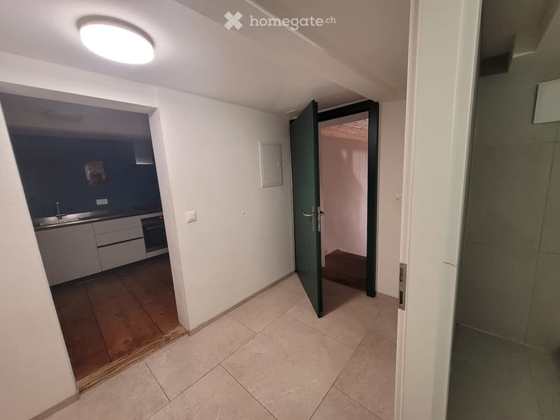 Empty hallway with tiled floor, white walls, and a round ceiling light. An open door leads to a kitchen area with white cabinets and a sink.
