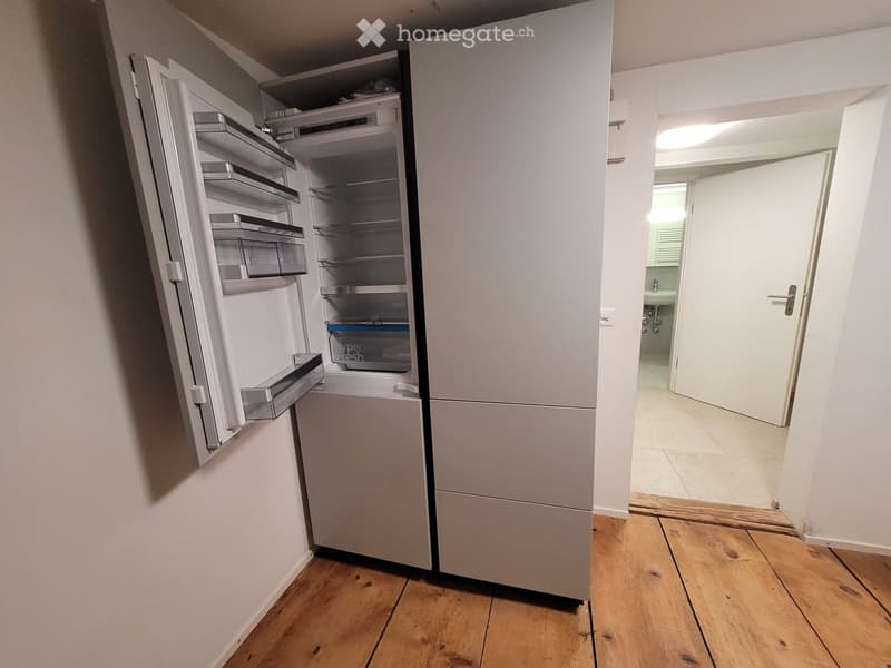 Empty room with wooden flooring, white walls, and an open refrigerator. A doorway leads to a bathroom with a sink and tiled floor.