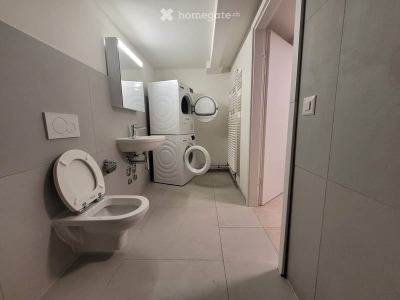 Modern bathroom with a toilet, sink, washing machine, mirror, and radiator. The space is clean and well-lit with tiled floors and walls.