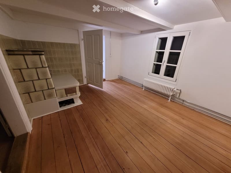 Empty room with wooden flooring, white walls, a window with white frame, a radiator, and a partially visible fireplace on the left side.
