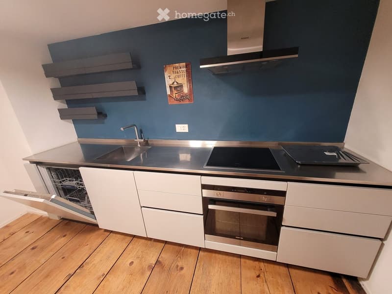 Modern kitchen with stainless steel appliances, white cabinets, and a dark blue accent wall. Features include a sink, faucet, dishwasher, oven, induction cooktop, and a kitchen island with built-in storage.