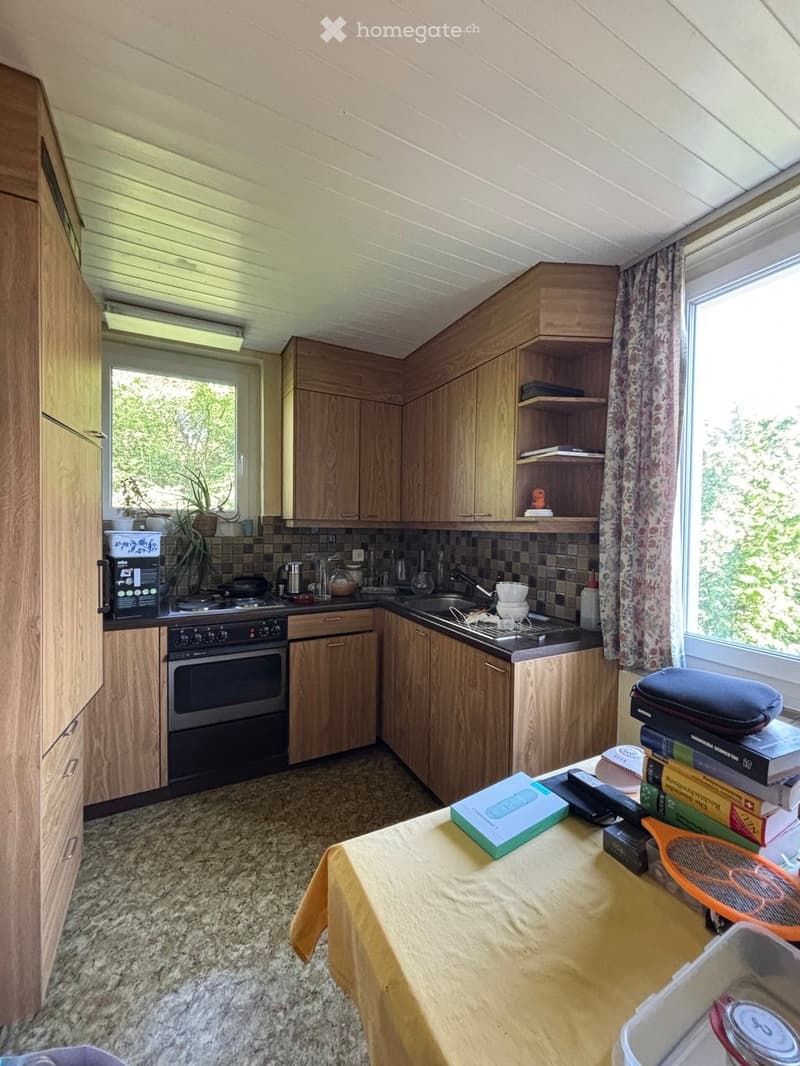 A cozy kitchen with wooden cabinets, a sink, stove, and oven. A small table with books, a remote control, and a tennis racket is visible, along with a potted plant on the windowsill.