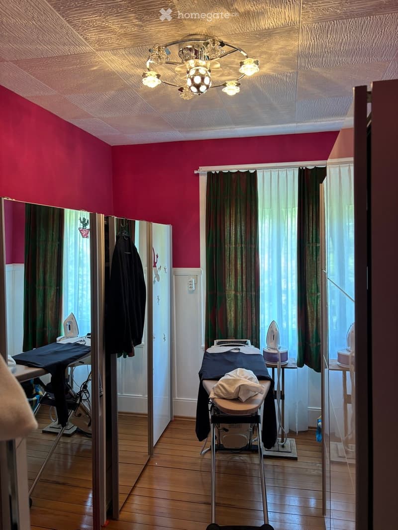 A well-lit room with wooden flooring, pink walls, and a chandelier. Contains an ironing board, iron, and a large mirror with a curtain.