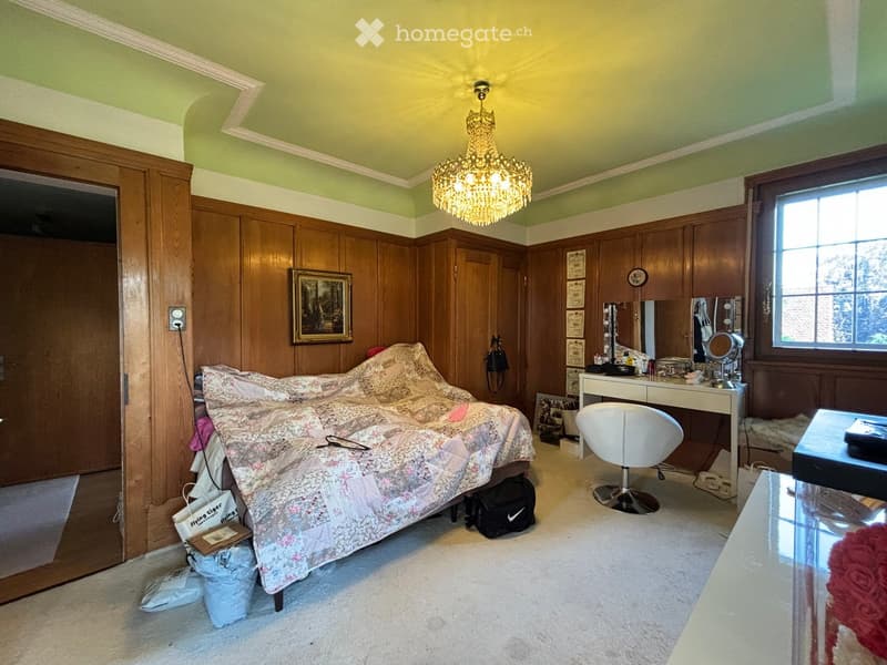 Bedroom with wooden panel walls, a chandelier, a bed with floral bedding, a vanity table with a mirror, a white chair, and a glass table.