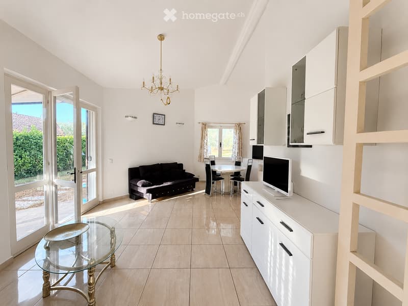 Bright living room with white walls, light wooden flooring, and large windows. Features a black sofa, glass coffee table, dining table with chairs, TV on a white cabinet, and a chandelier.