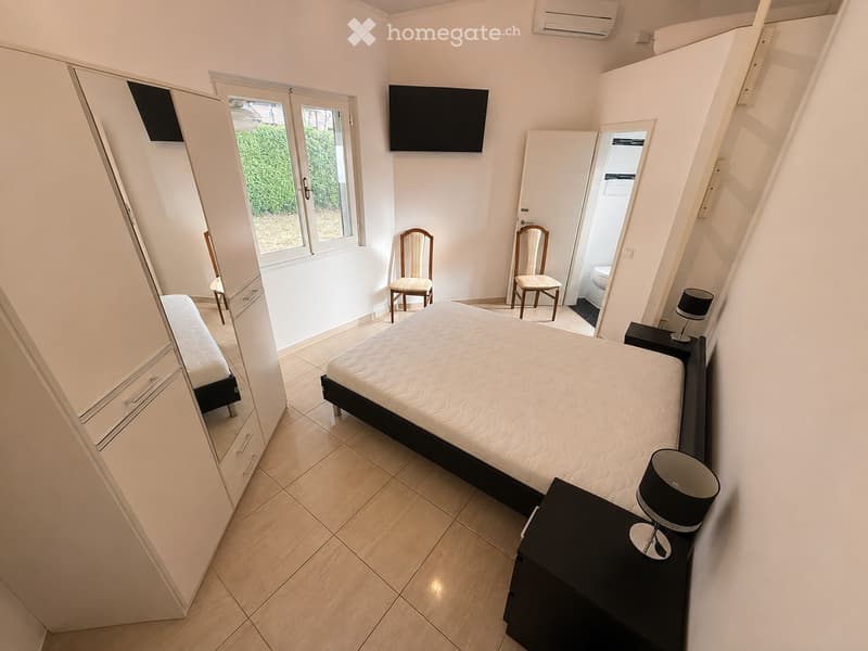 Modern bedroom with a double bed, nightstand with lamp, two chairs, a large mirror, and a television mounted on the wall. The room features tiled flooring and has a sliding glass door leading to a balcony.