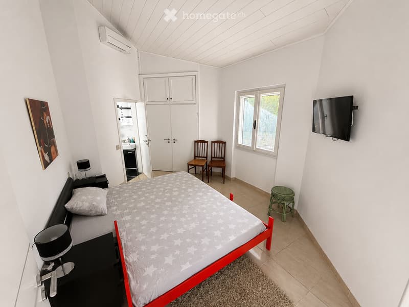Bright bedroom with a red-framed bed, nightstand, lamp, two wooden chairs, green stool, mirror, cabinet, and television mounted on the wall.