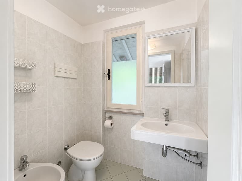 Modern bathroom with beige tiled walls and floor, featuring a toilet, sink with faucet, mirror, and a window with frosted glass.