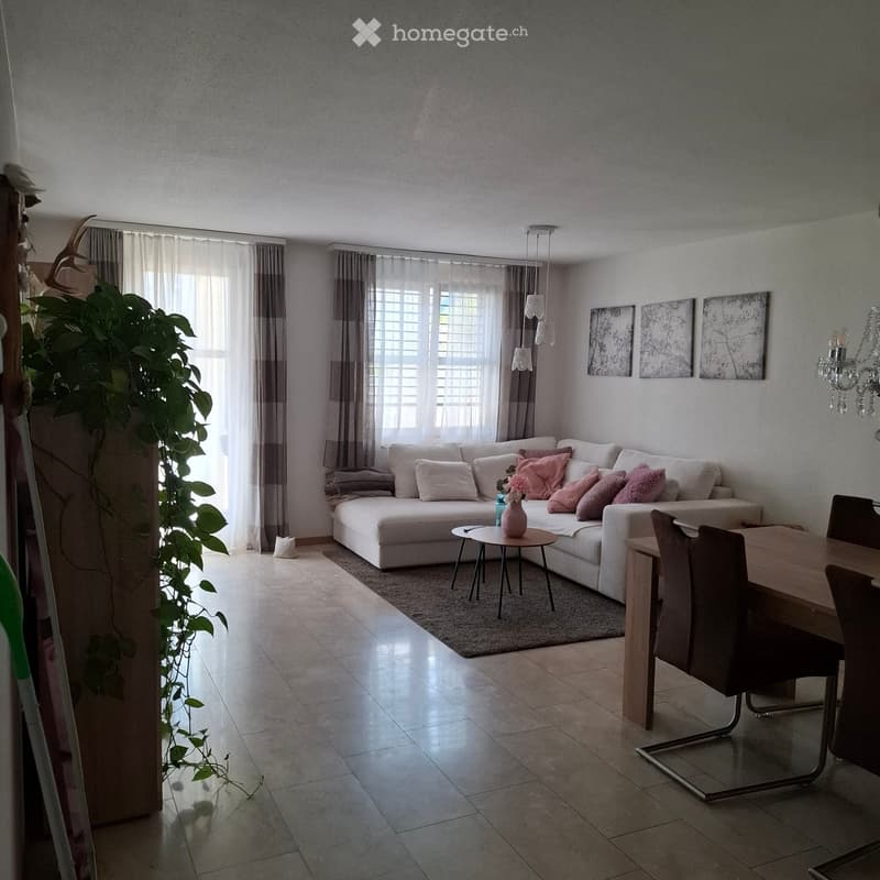 Modern living room with a white sofa, pink cushions, a small round coffee table, a dining area with a wooden table and chairs, and large windows with curtains.