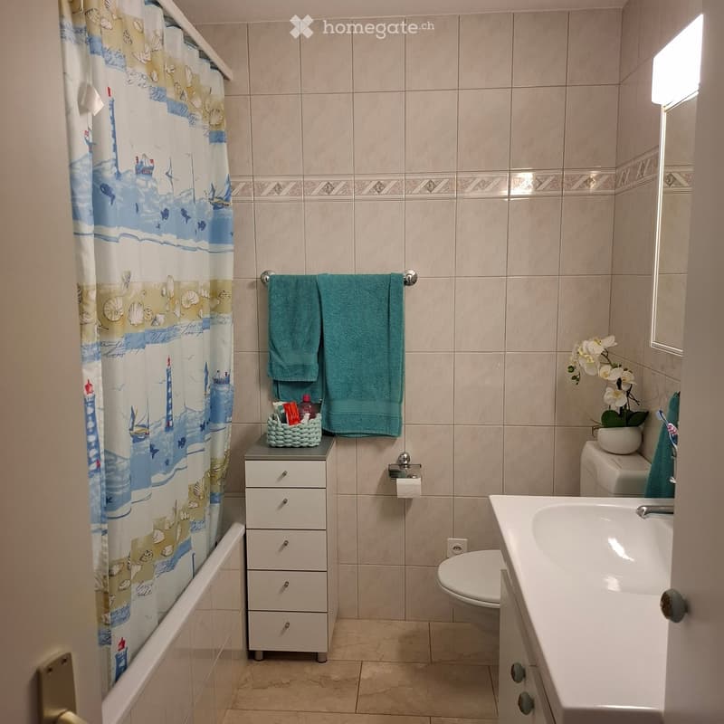 Bathroom with beige tiled walls and floor, bathtub with a colorful shower curtain, toilet, sink, towel rack with green towels, small basket with toiletries, and a flower pot on the toilet tank.