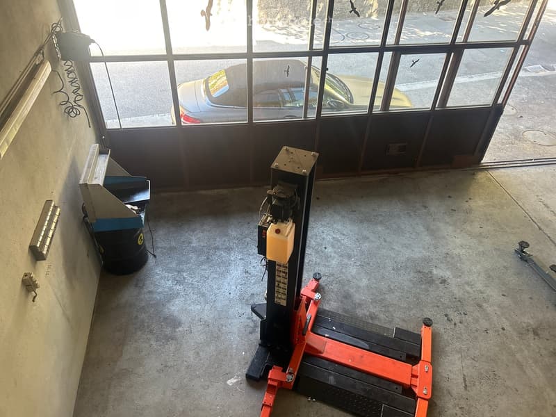 Empty garage with concrete floor, large glass door showing a car outside, and a few pieces of equipment including a hydraulic jack and a black container.