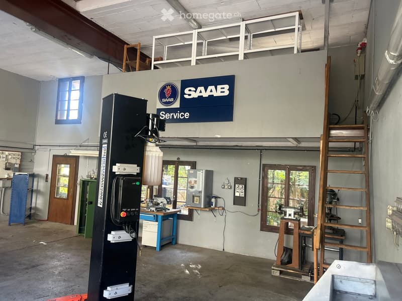 Workshop space with a large SAAB sign, various tools and equipment, a staircase leading to an upper level, and multiple stools.