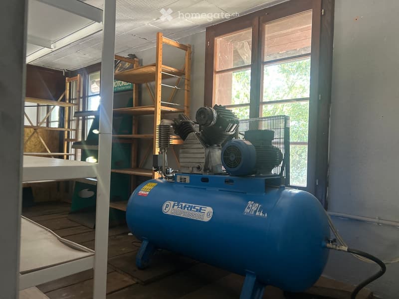 A workshop or storage room with a large blue air compressor on the floor, a wooden shelf unit, and a window letting in natural light.