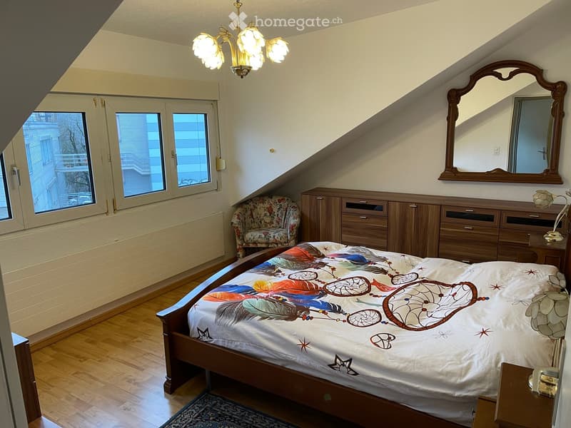 A bedroom with wooden floor, a bed, wooden wardrobe, wooden cabinet, a bedspread with a dreamcatcher print, mirror, two windows, chandelier, wooden nightstand with a lamp, a floral chair