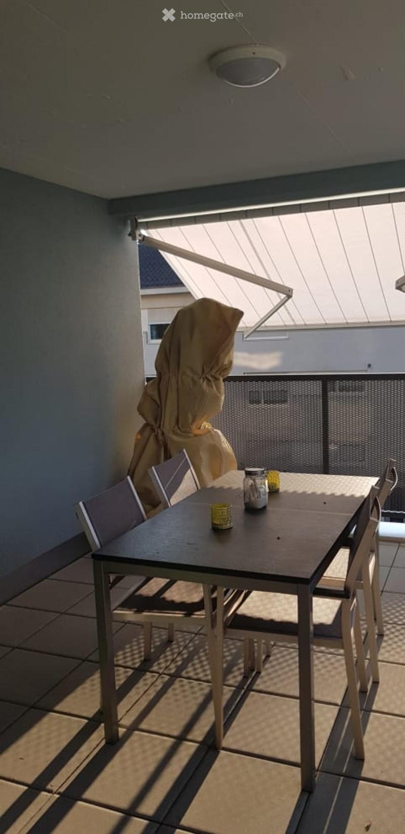 Balcony with a dining table, chairs, and a covered umbrella. The space features a retractable awning and a tiled floor.