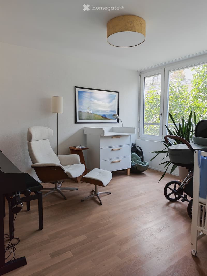 A well-lit room with wooden flooring, a large window, a white cabinet, a comfortable chair, a small table, a lamp, a picture frame, and a baby stroller.