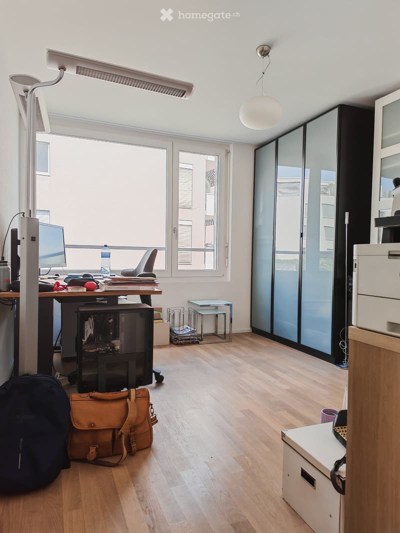 Bright, modern office space with wooden flooring, large windows, a desk with a computer, a chair, and various office supplies.