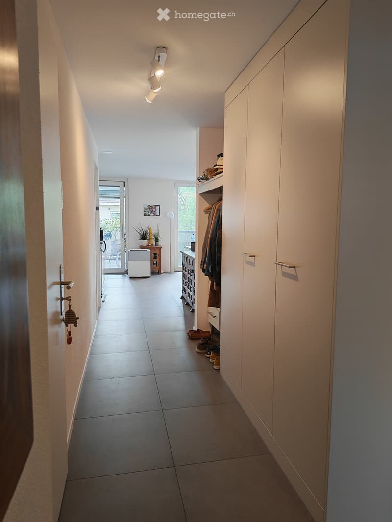 A modern hallway with tiled flooring, white walls, built-in storage cabinets, and a small shoe rack with shoes and coats. The hallway leads to an open area with a small table, plants, and a view of the outdoor space.