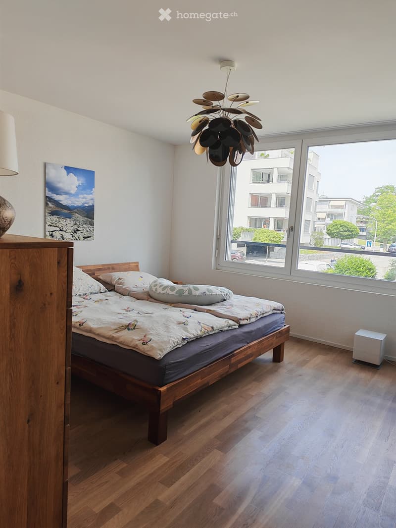 A modern bedroom with a wooden bed, colorful bedding, a nightstand with a lamp, and a large window offering a view of the city.