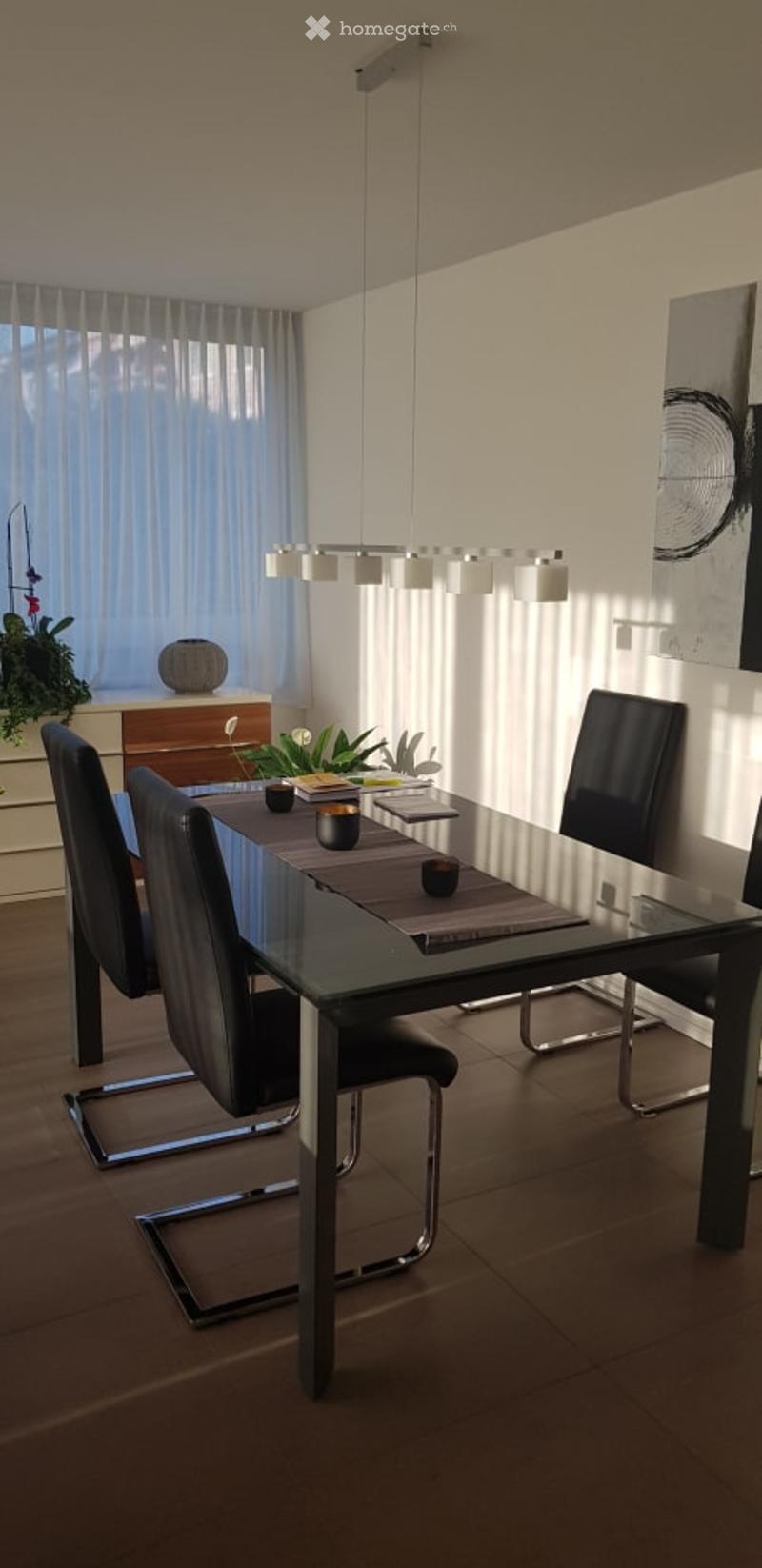 Modern dining area with a sleek black glass dining table, four black chairs, pendant lighting, a small cabinet with a plant, and artwork on the wall.