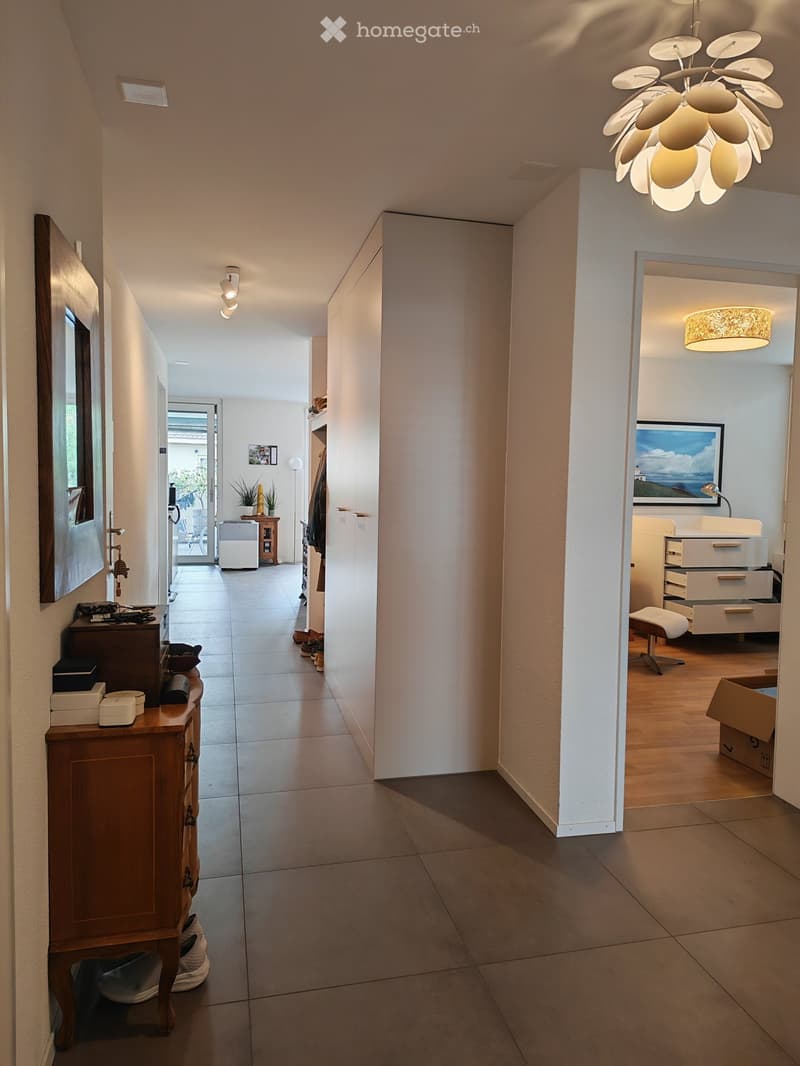 Bright hallway with tiled floor, white walls, and modern lighting. Features a wooden console table with various items, a large mirror, and an open doorway leading to another room with wooden flooring and furniture.
